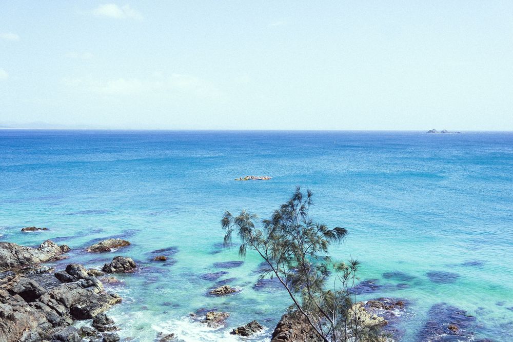 Panoramic view of turquoise ocean waves crashing against rocky coastline with tropical vegetation and clear blue sky.