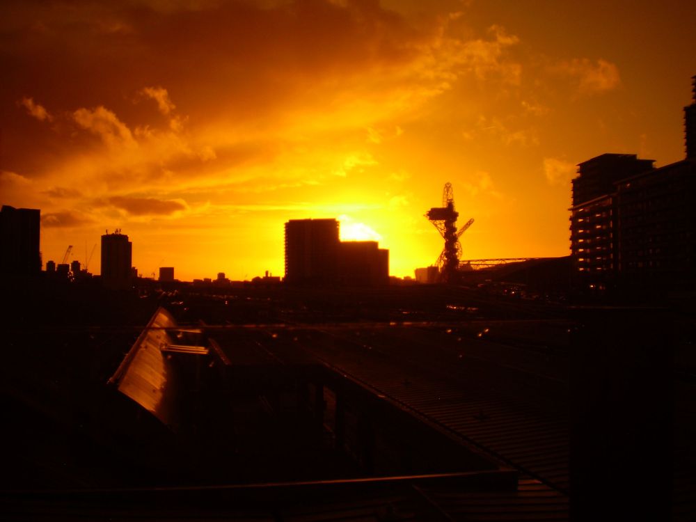 Dramatic orange sunset silhouettes city buildings and an industrial structure against a vivid golden sky.