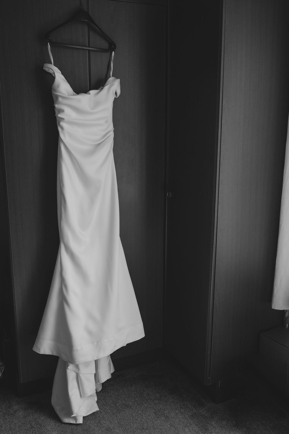 A classic white wedding dress with a mermaid silhouette hangs elegantly on a black hanger in a dark room.