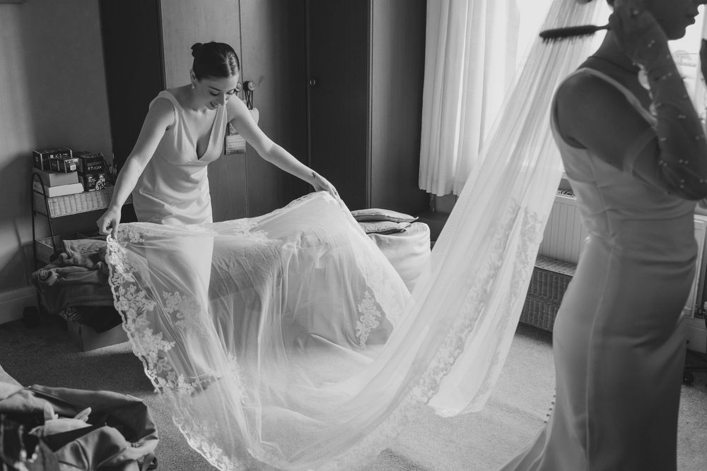 Two figures help arrange a flowing lace wedding veil in a sunlit room during wedding preparations.