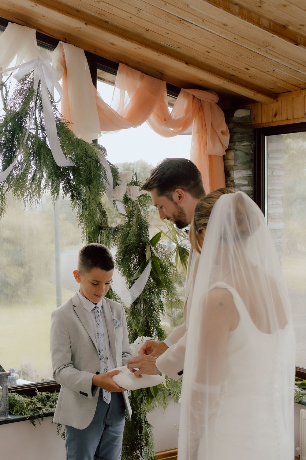 A wedding ceremony in a rustic venue decorated with evergreen branches and peach-colored draping.
