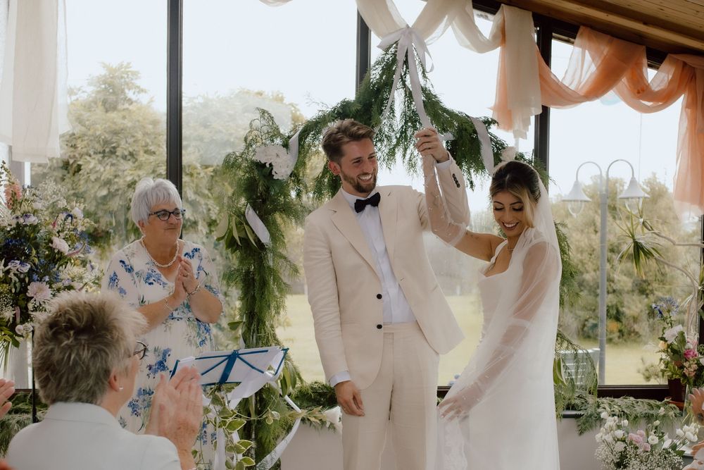 A joyous wedding ceremony takes place under a decorated gazebo with greenery and flowing white fabric.