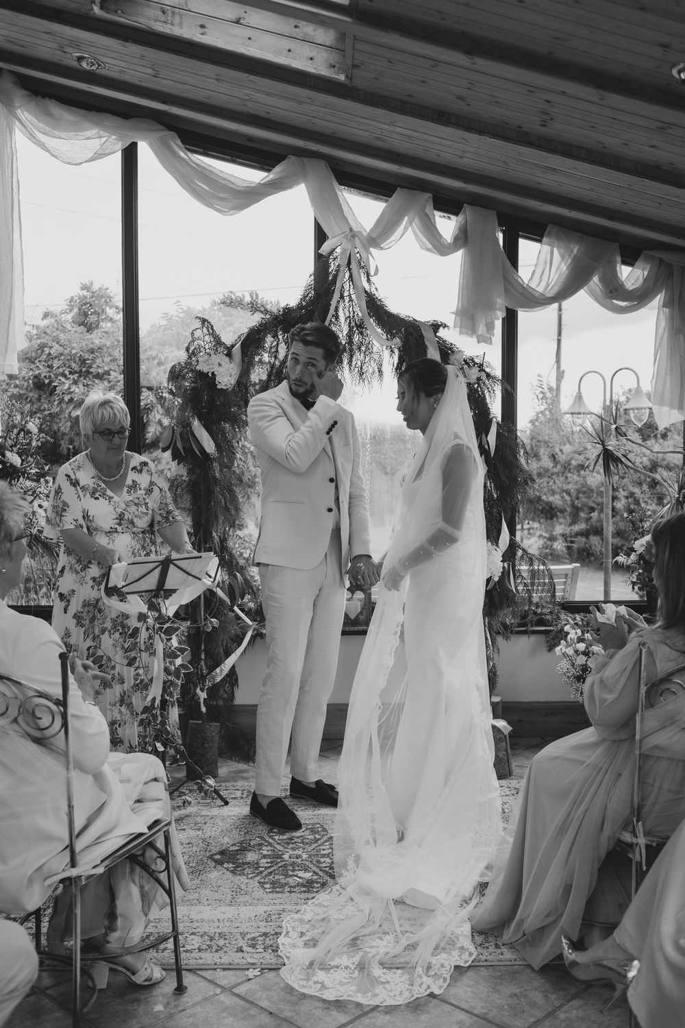 A black and white photograph of a wedding ceremony under a decorated canopy with flowing white drapes and floral arrangements.