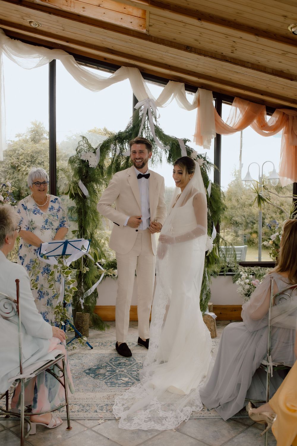 An intimate wedding ceremony takes place under a draped canopy with greenery and white fabric decorations.