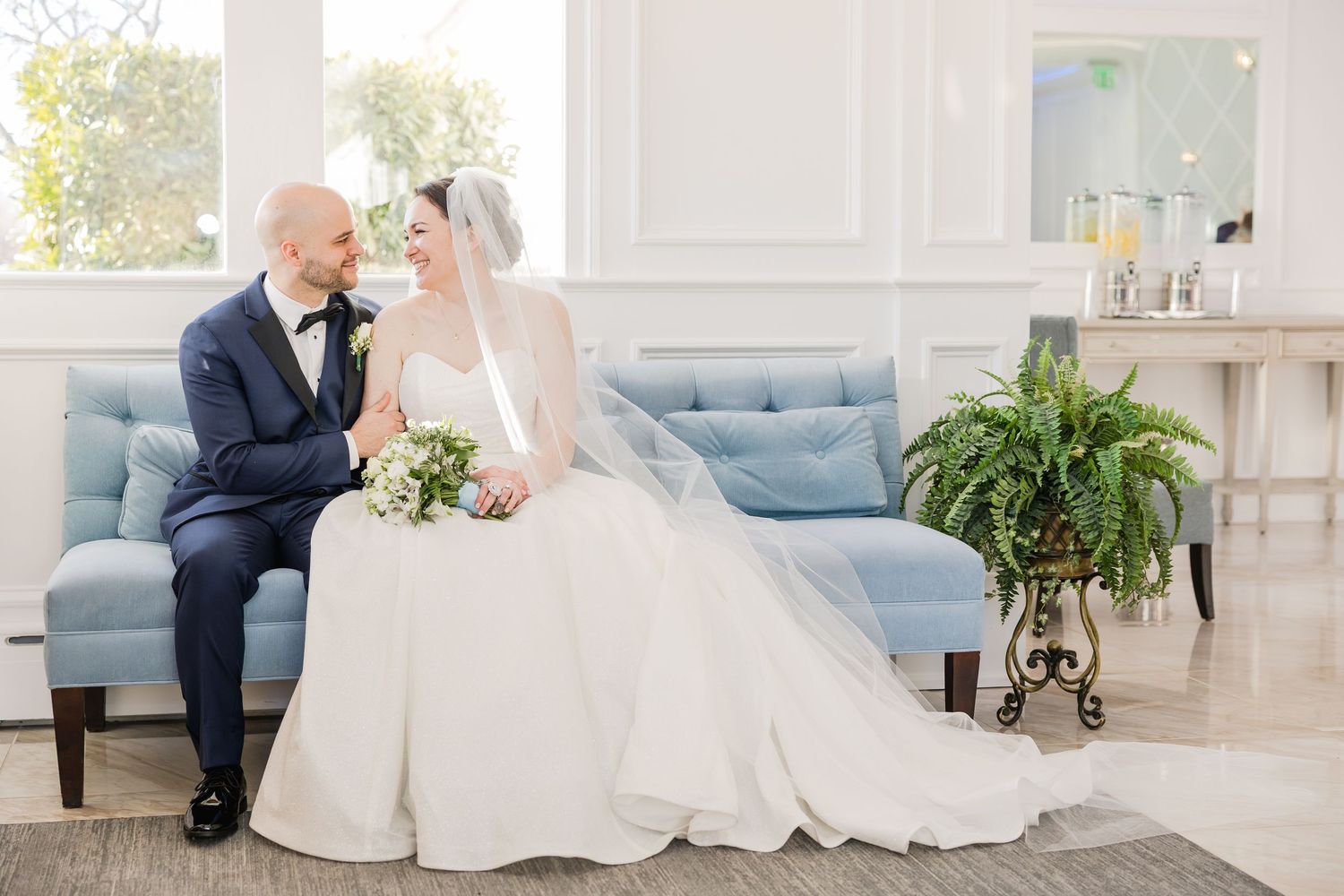 Wedding couple poses on light blue furniture in an elegant white interior.