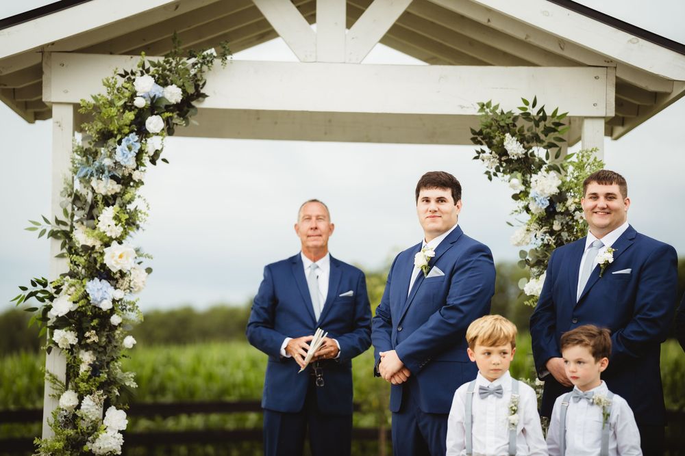 Wedding party stands by floral arch decorated with white roses and greenery at an outdoor ceremony.