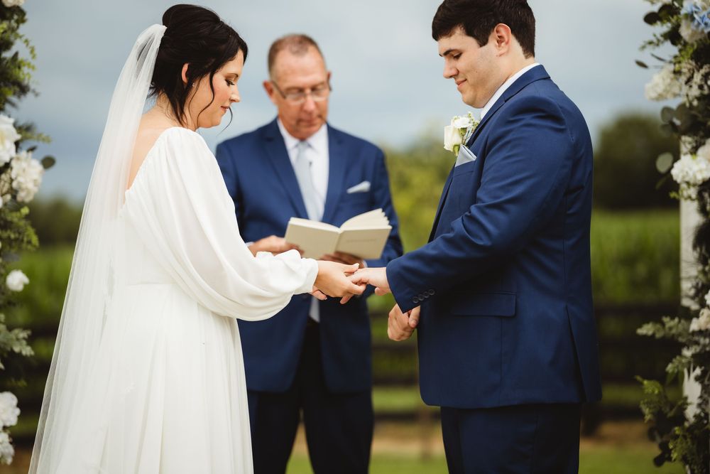 A wedding ceremony takes place outdoors with a couple exchanging rings in front of an officiant beneath a cloudy sky.
