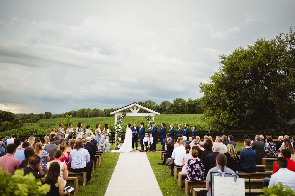 An outdoor wedding ceremony with white chairs and an arbor set against rolling green hills on a cloudy day.