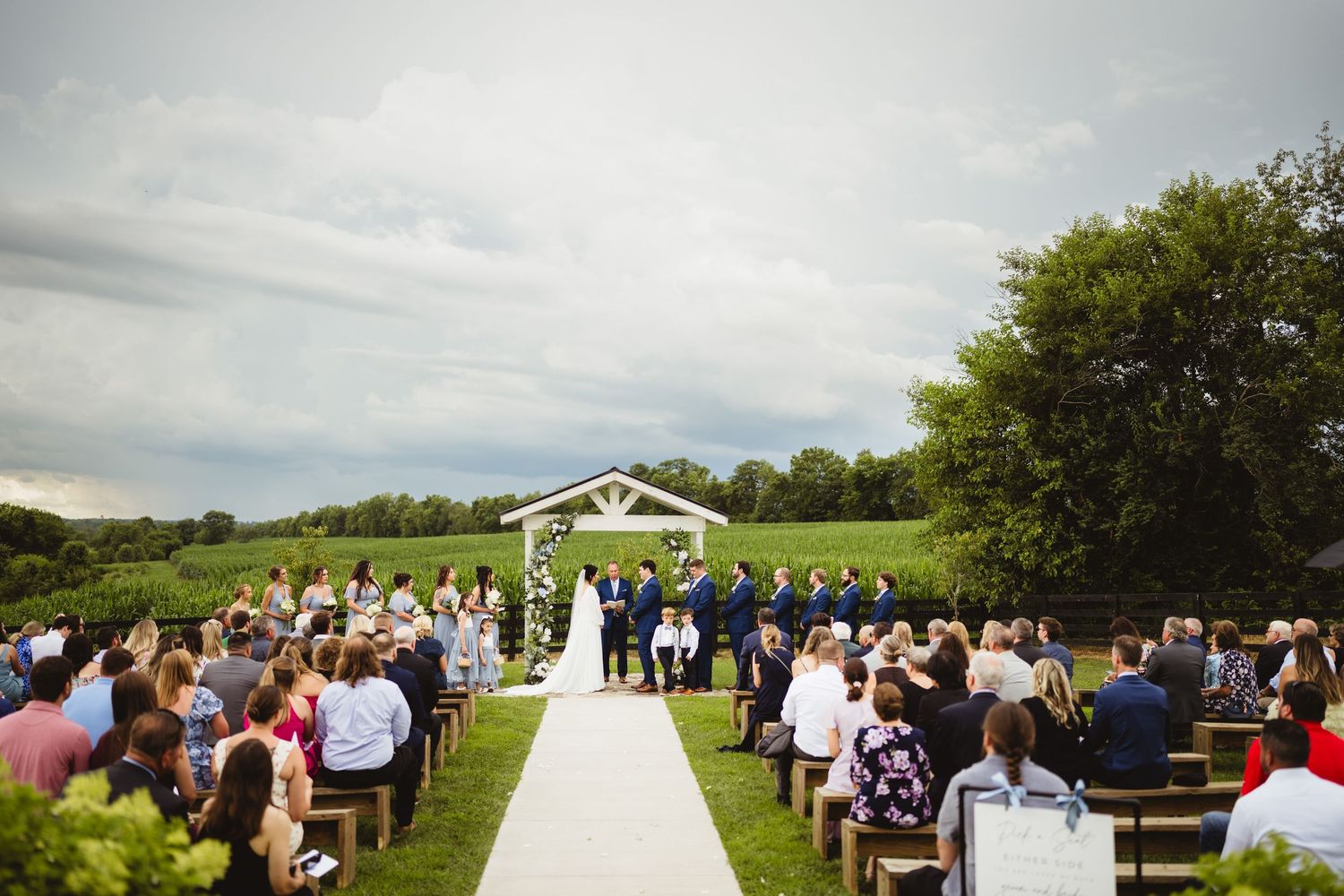 An outdoor wedding ceremony with white chairs and an arbor set against rolling green hills on a cloudy day.