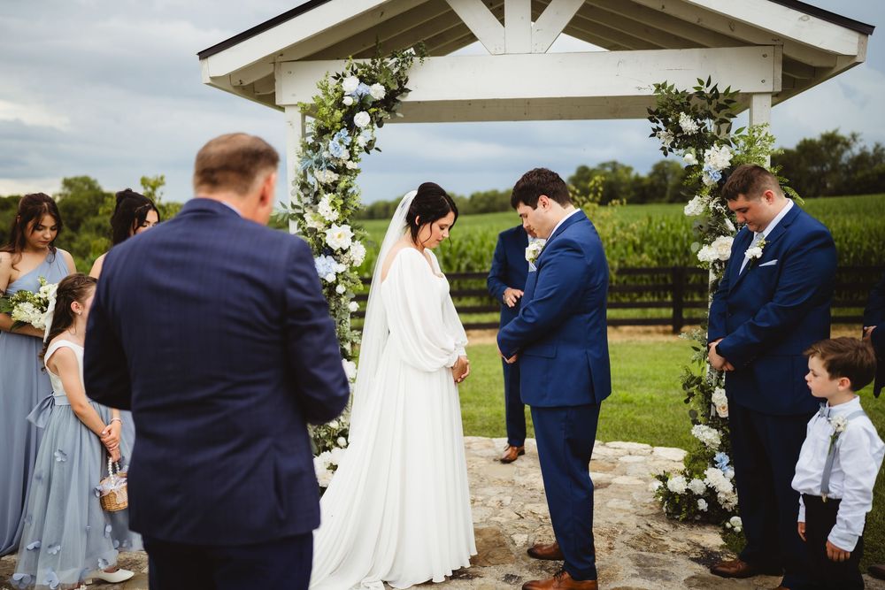 Wedding party gathered outdoors under a white beam gazebo structure during a countryside ceremony.