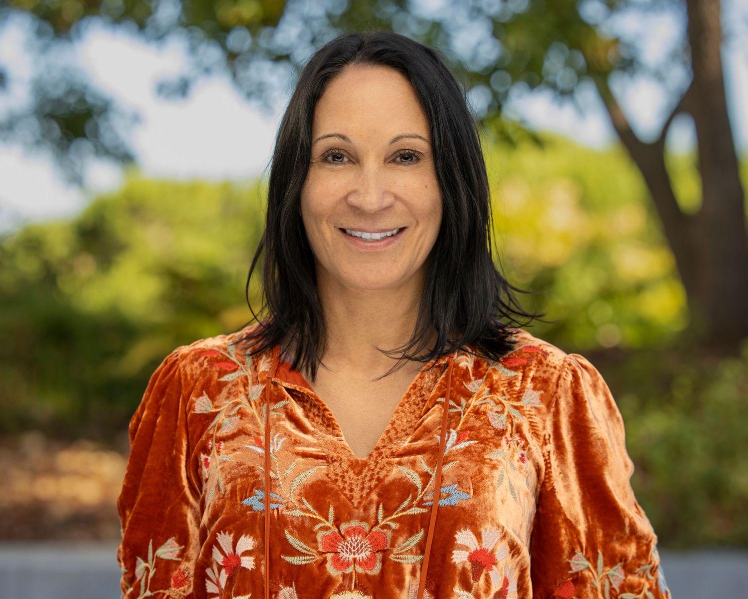 A person wearing an orange floral blouse poses outdoors on a sunny day with trees in the background.