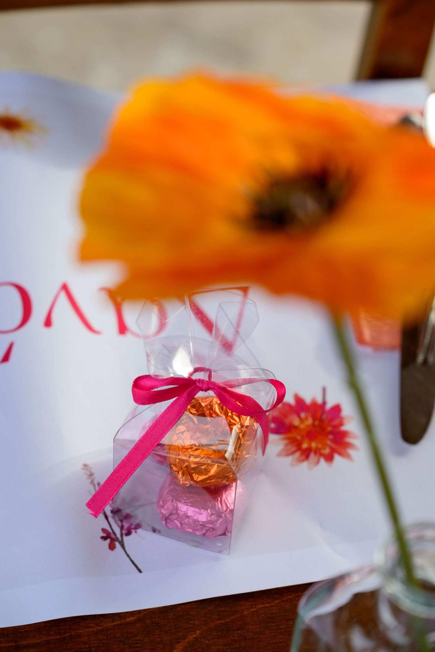 A small pink favor box with ribbon sits beside an orange flower on a white surface.