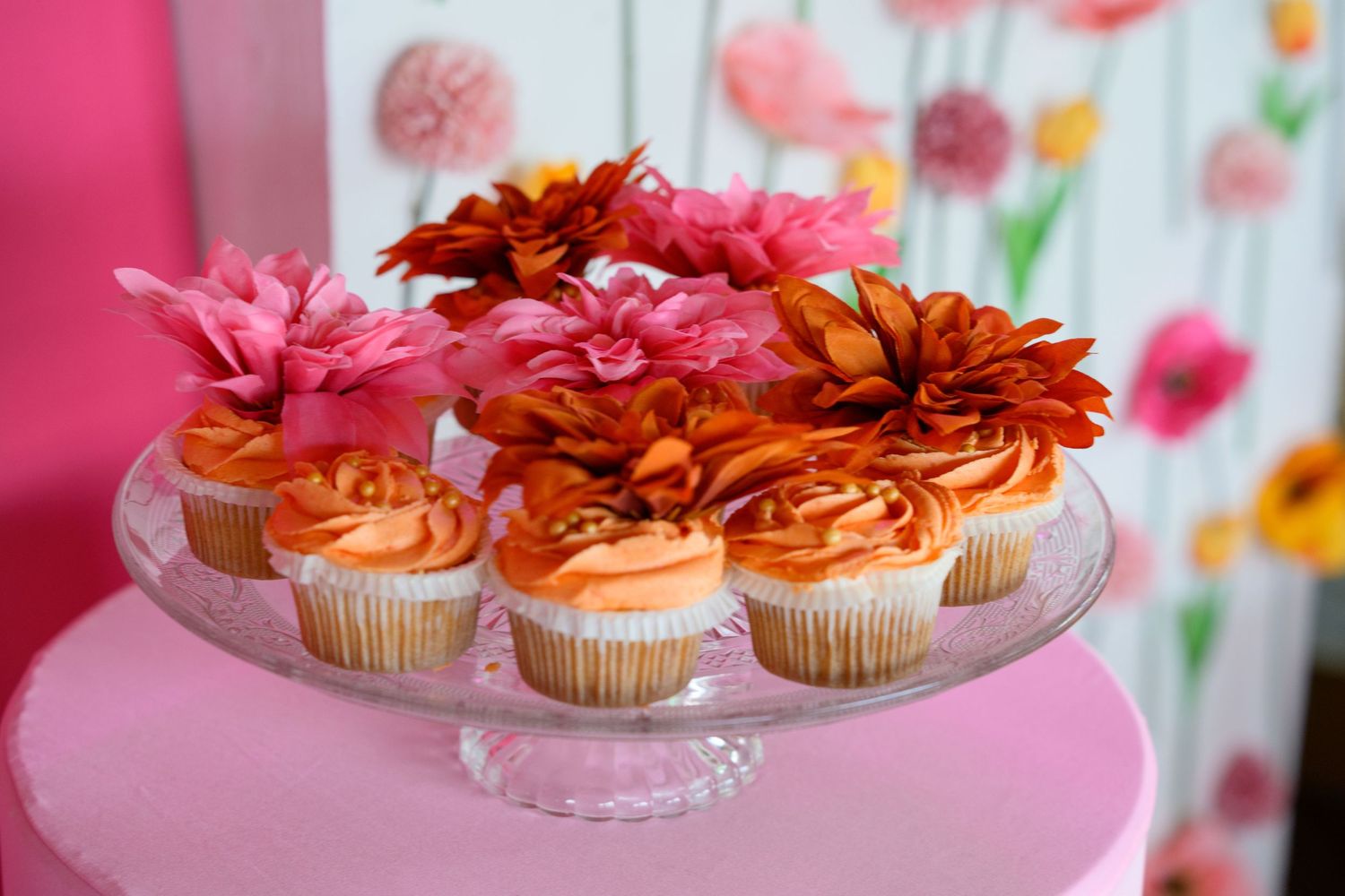 Pink and orange cupcakes topped with flowers displayed on a glass cake stand against a floral backdrop.