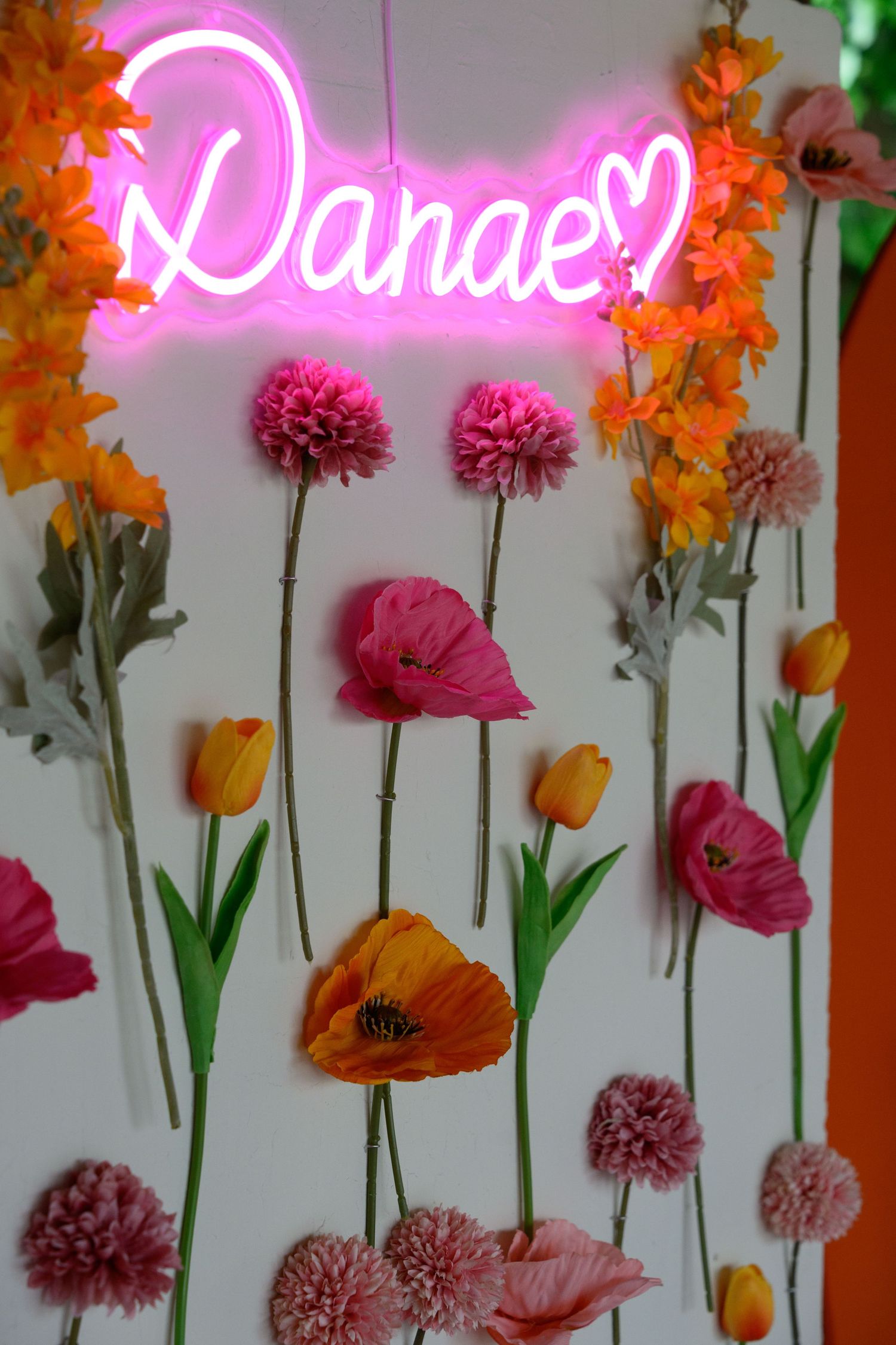 Pink neon 'Danae' sign displayed against white wall with colorful flowers including orange snapdragons and pink poppies.