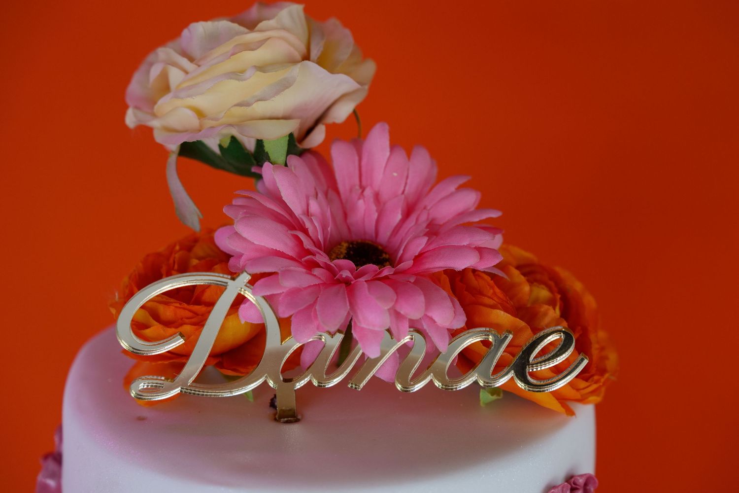 White celebration cake decorated with pink gerbera daisies and cream roses with metallic 'Danae' topper.