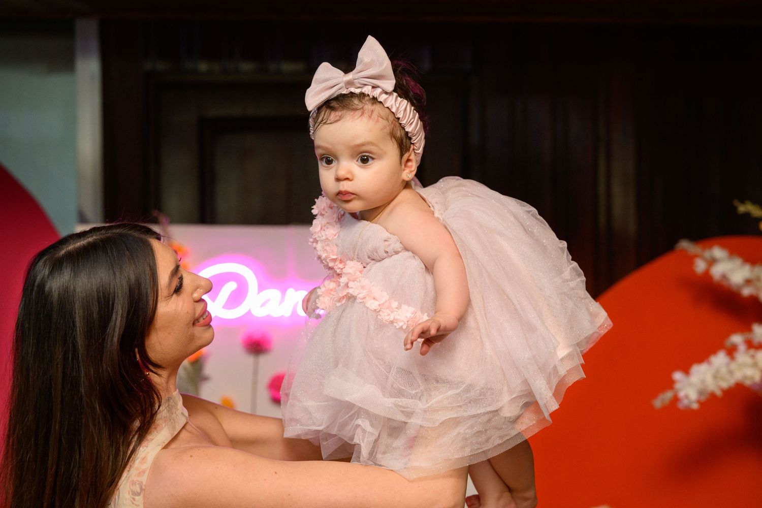 Baby in tulle dress and bow headband posed against floral backdrop with neon sign.
