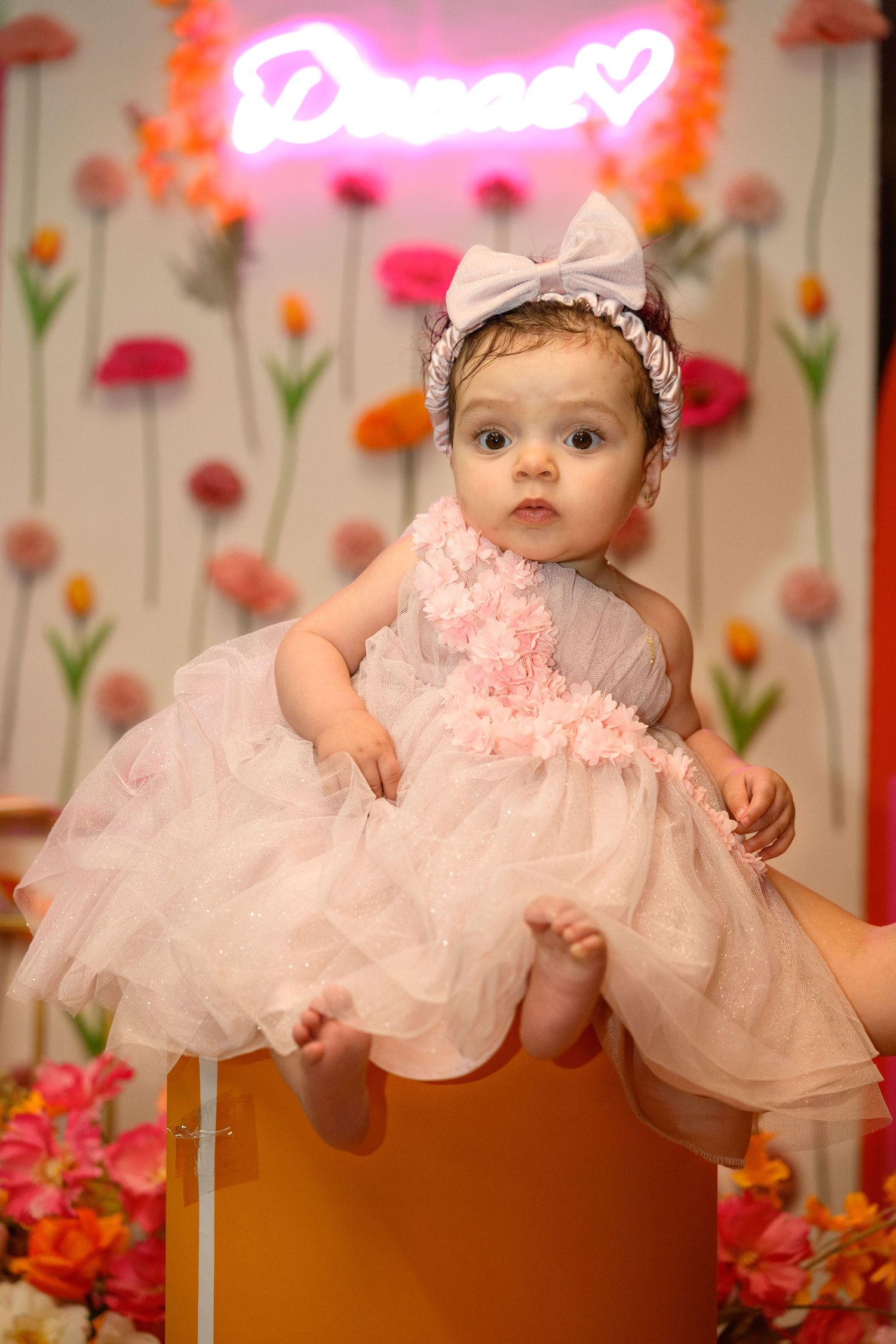 Baby wearing pink tulle dress and bow against floral wall with neon signage.