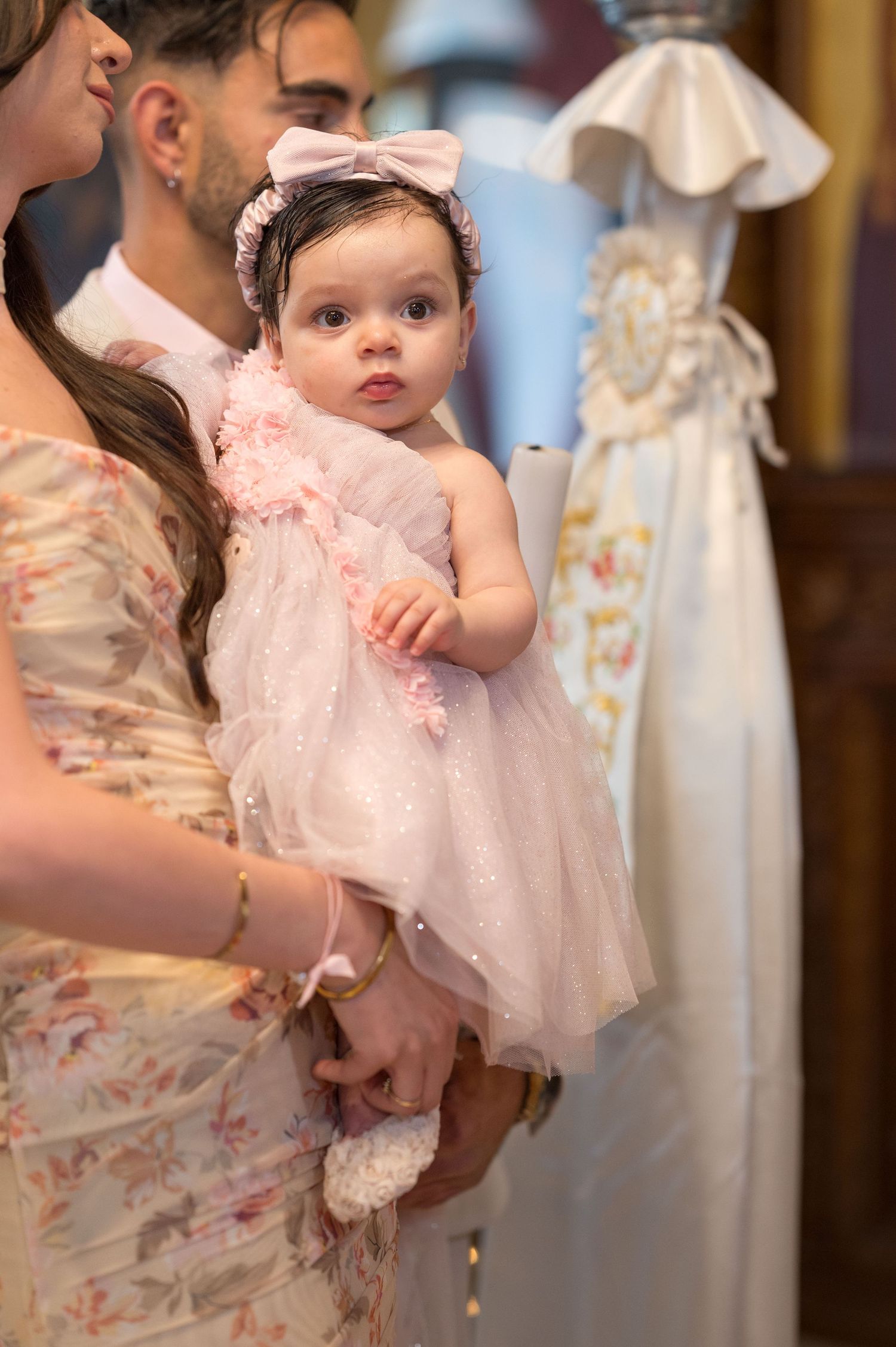 Infant wearing a frilly white and pink dress at a formal event.