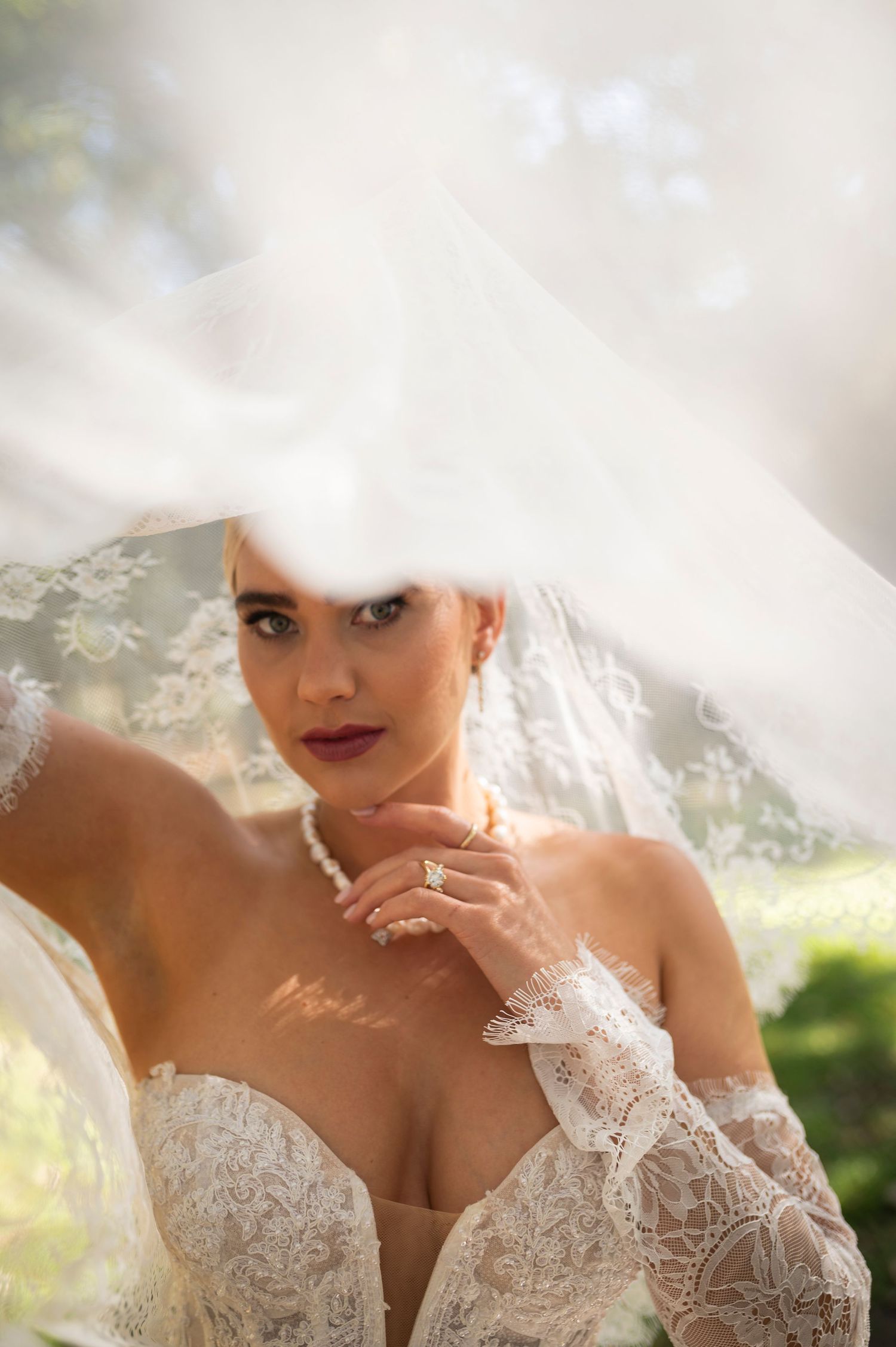 A bride poses in a delicate white lace wedding gown with off-shoulder sleeves and veil in soft natural lighting.
