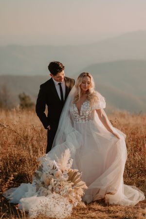 Alissa in white dress and Andrew in black suit stand together in an open field with mountain views.