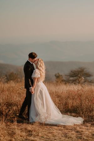Alissa and Andrew, both in extravagant wedding attire, share a kiss in a field with mountains in the background.
