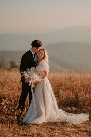A couple in wedding attire embraces in a field with mountains in the background during sunset.