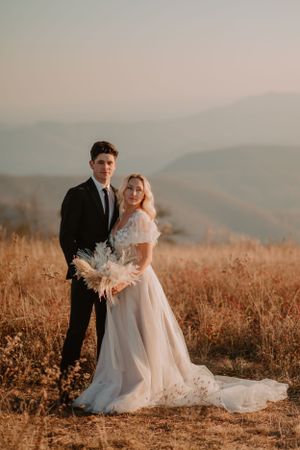Alissa and Andrew stand in a grassy field with mountains in the background, dressed in wedding attire.
