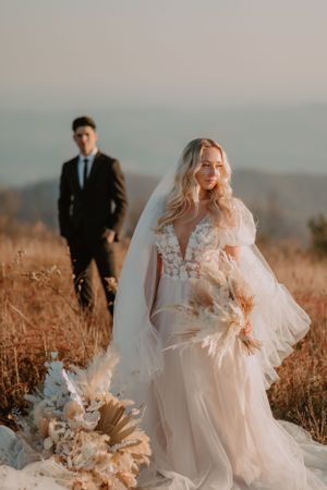 Alissa stands in her wedding dress holding her bouquet in a field with Andrew in a black suit in the background.
