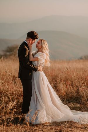 Alissa and Andrew embrace in a grassy field with mountains in the background during sunset.