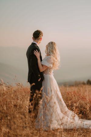 Alissa and Andrew stand in a field at sunset, facing away and gazing at the distant horizon.