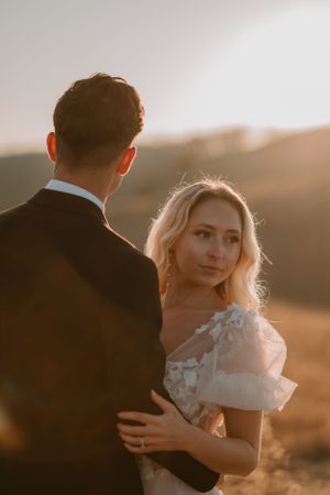 Bride and groom stand close together in a sunny outdoor setting, with Alissa looking to the side and Andrew facing away.