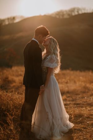 Alissa and Andrew lean in for their first kiss in a sunlit field with hills in the background.