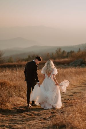 Alissa and Andrew hold hands and walk into the sunset after their elopement ceremony, groom carrying his bride's dress.