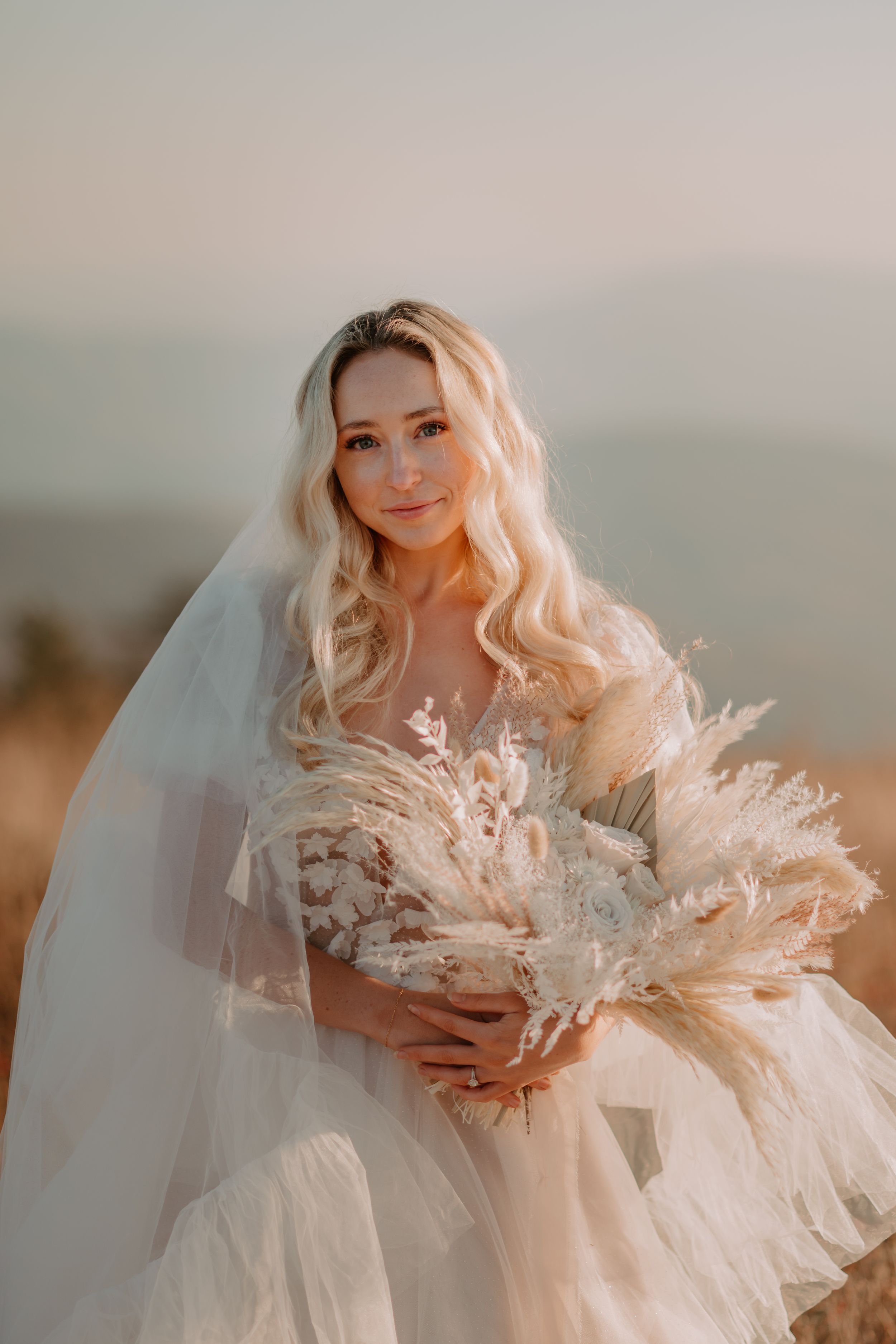 Alissa, a bride with long blonde hair, holds a bouquet of dried flowers, standing outdoors with a soft focus background.
