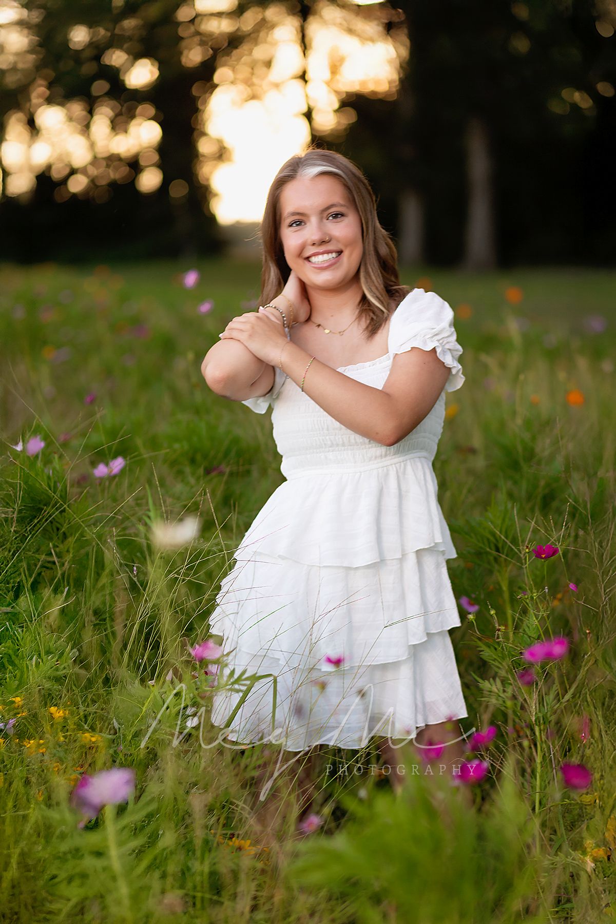 A teen in a white dress poses in a wildflower field at sunset with soft lighting in the background. MegMc Photography, senior pictures
