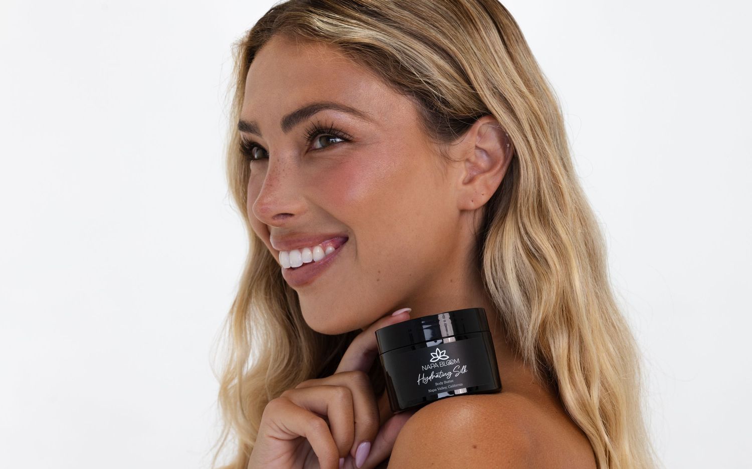 A woman with blonde hair smiles while holding a black cosmetic jar against a white background.