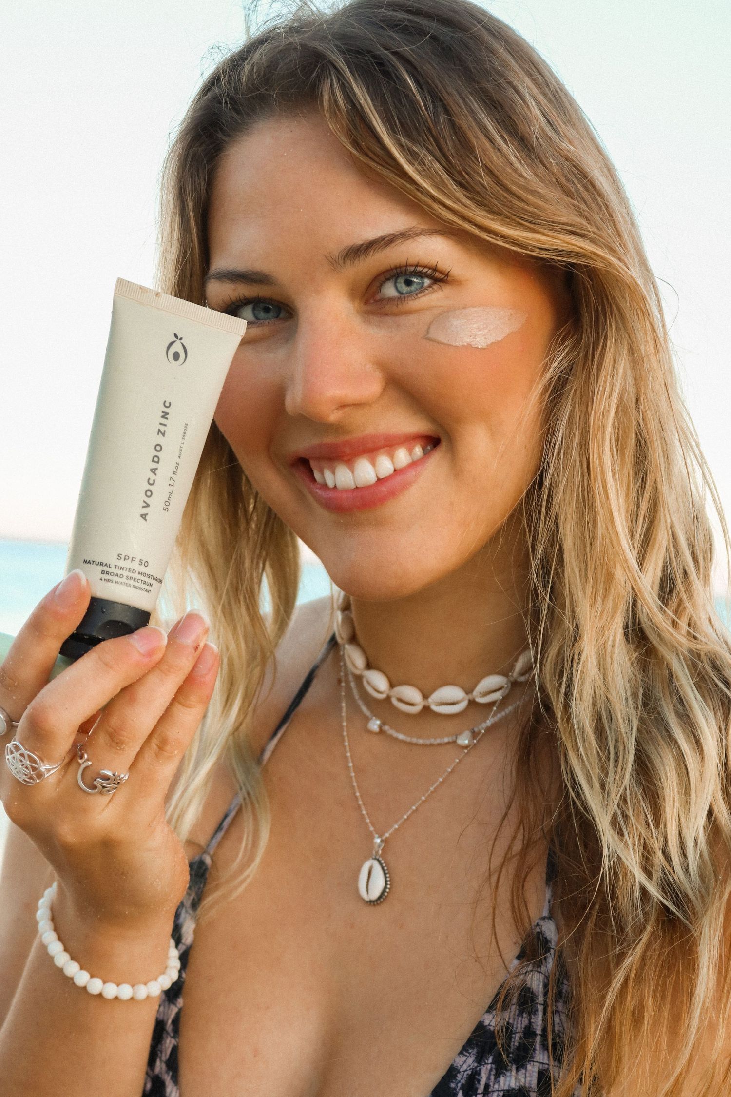 Woman displaying skincare product while wearing beach jewelry and summer attire.