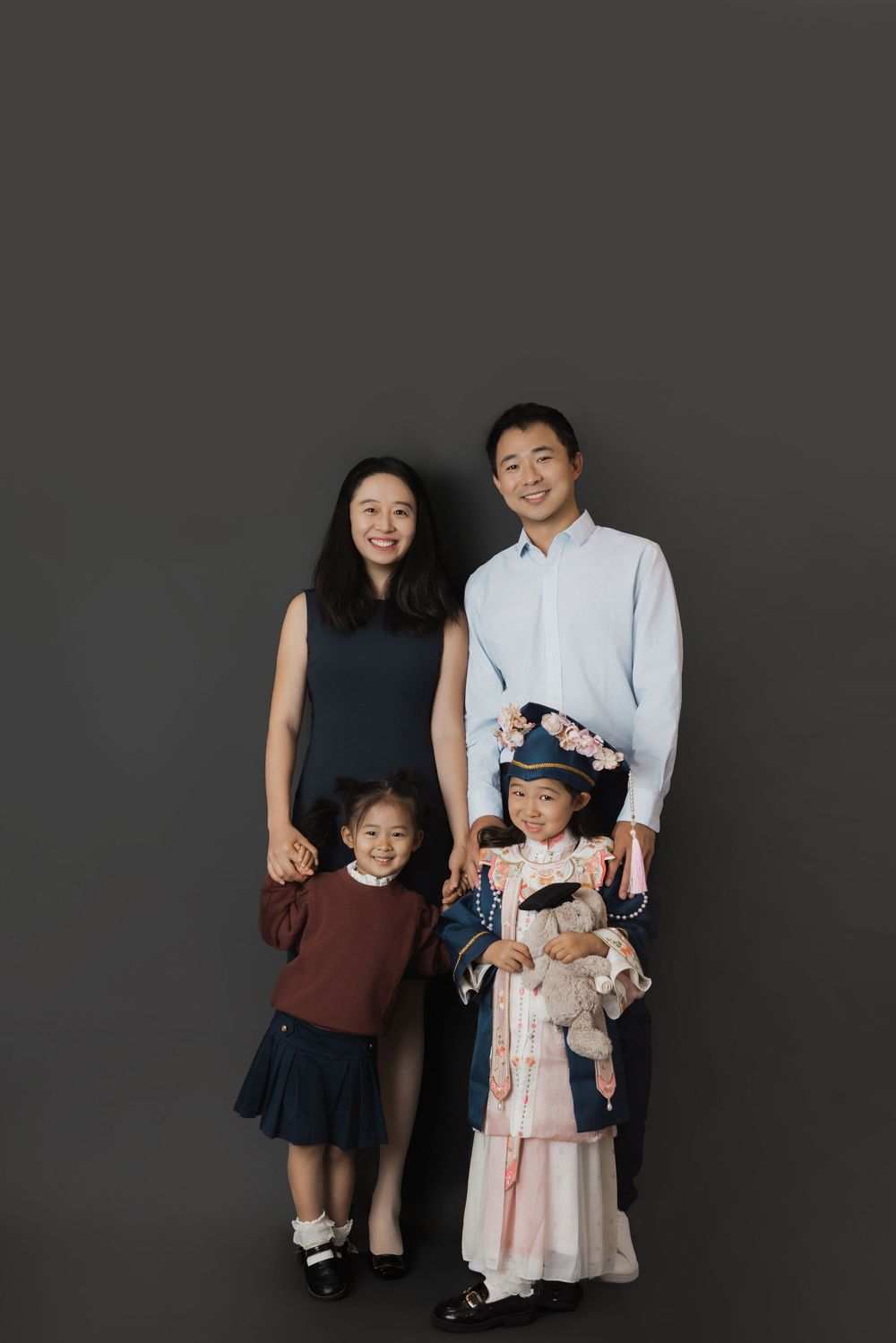 Family of four posing for a portrait, parents with two young daughters, one in traditional dress, against gray background.