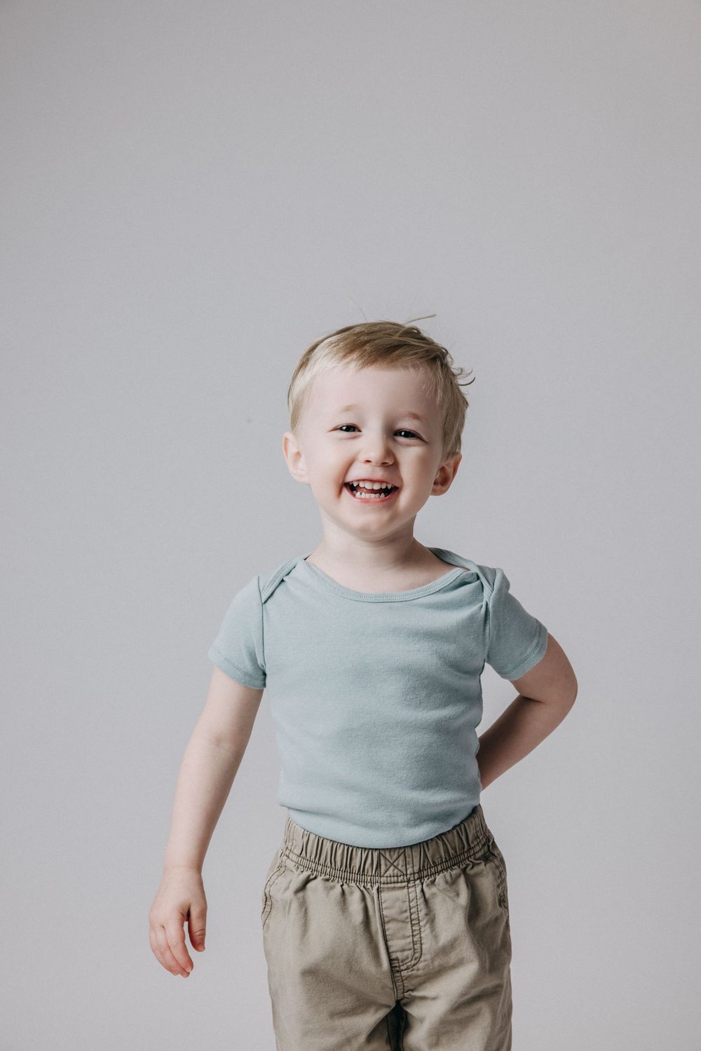 Toddler in light blue shirt smiles joyfully against grey background.