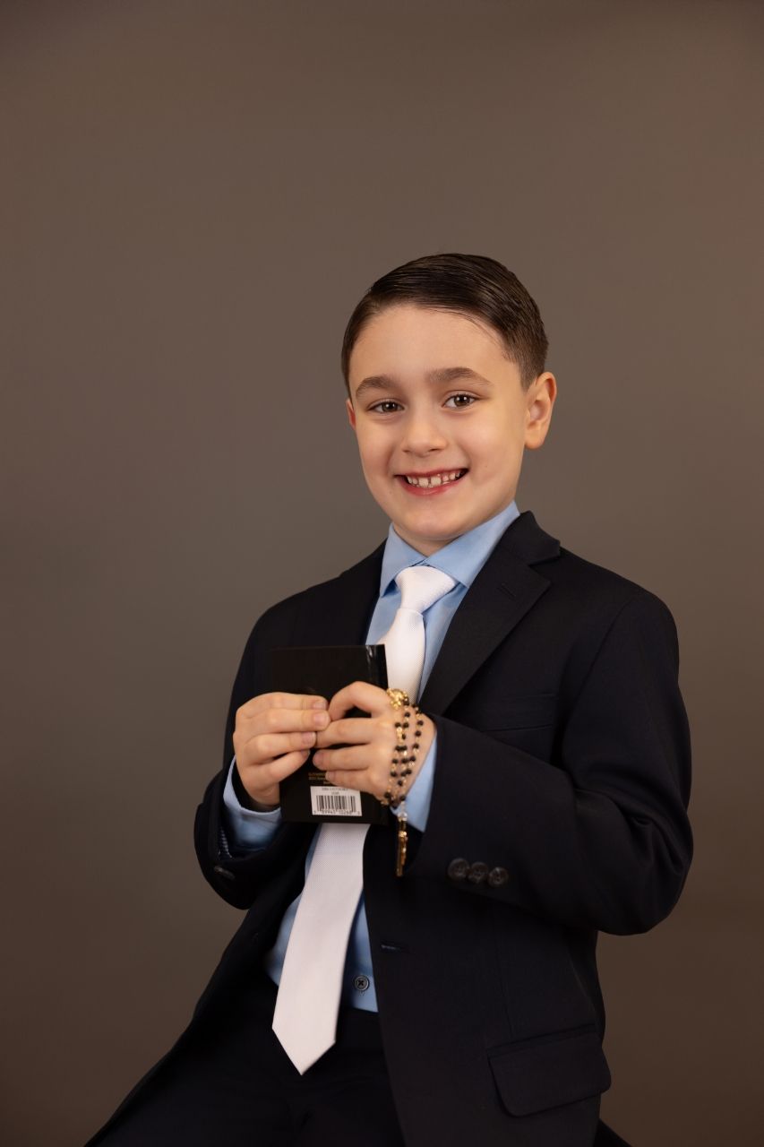 Young student in a formal black suit and light blue tie smiles while holding a small object against brown background.