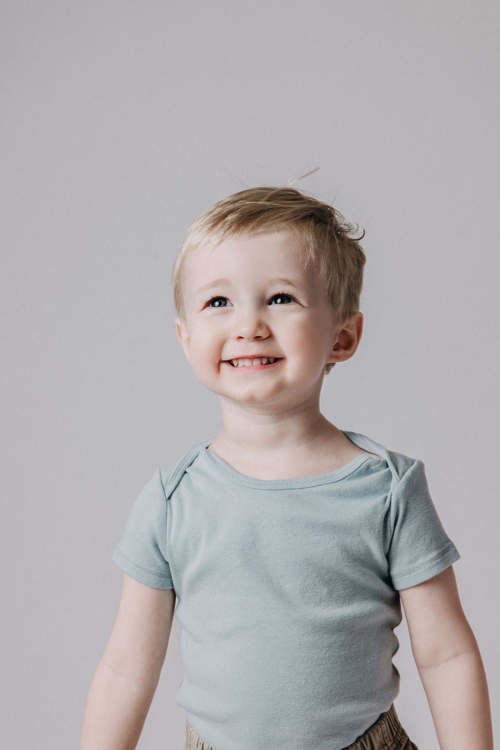 Toddler in light blue shirt looks up with bright smile against grey background.