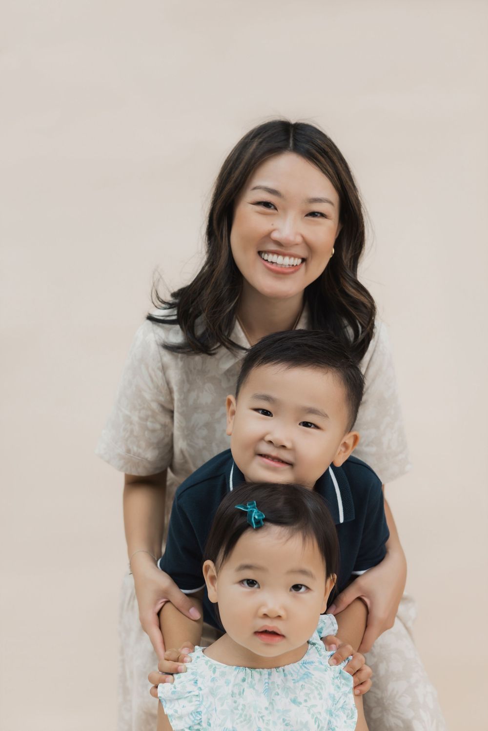 Smiling Asian mother posing with two young children against a beige background in a warm family portrait.