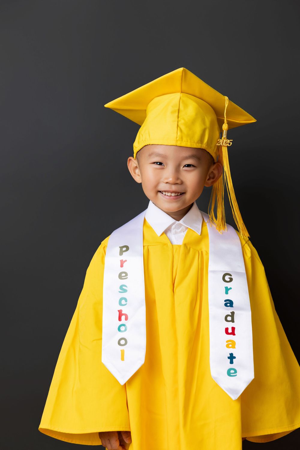 Young graduate in bright yellow cap and gown with decorated white sash stands against dark background.