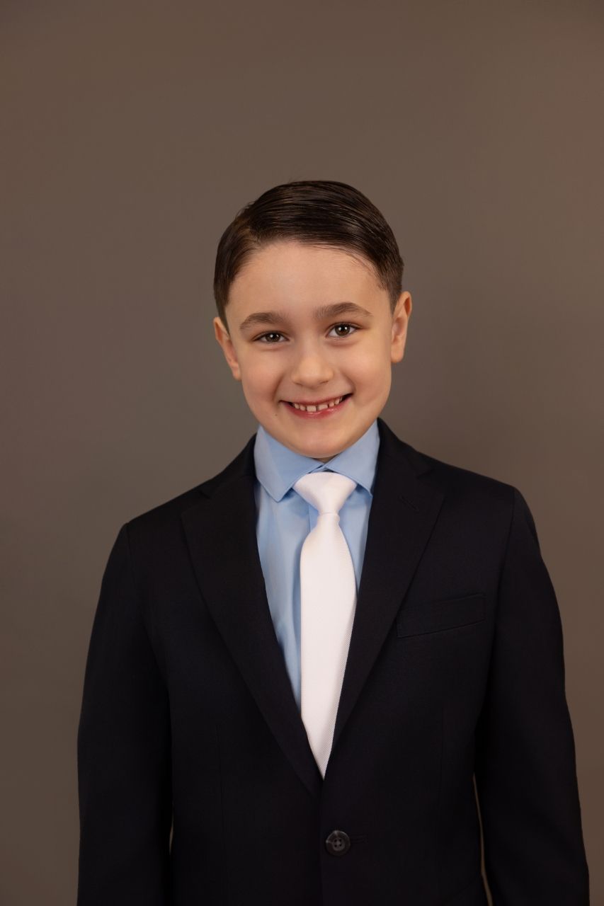 Young child wearing a formal black suit with light blue tie smiling against brown studio backdrop.