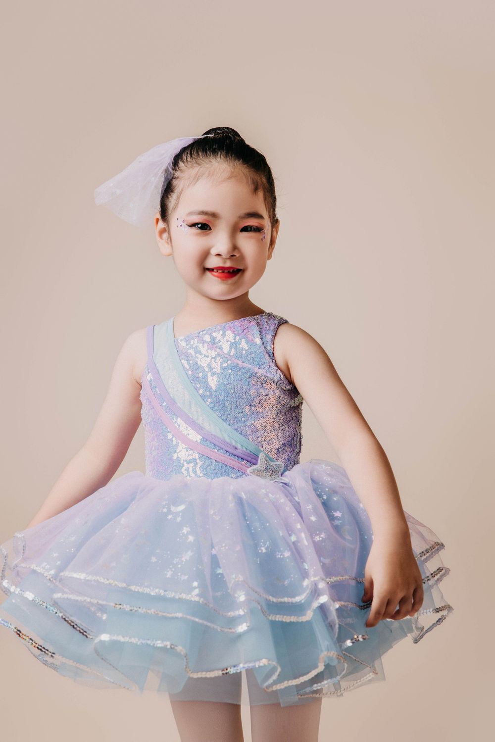 Young dancer poses in sparkly blue and pink tutu dress against neutral background.