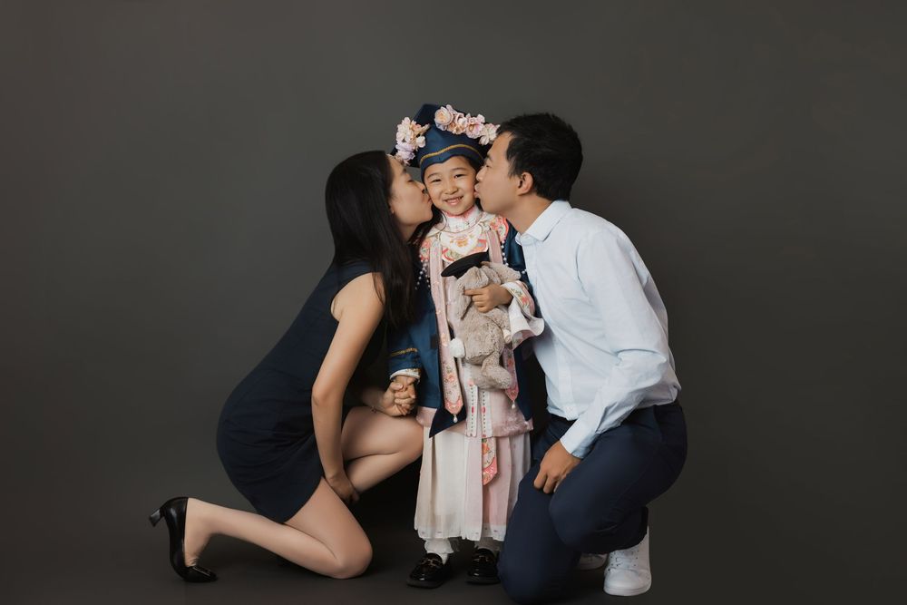 A young girl in traditional attire and floral crown receives kisses from her proud parents during a studio portrait session.