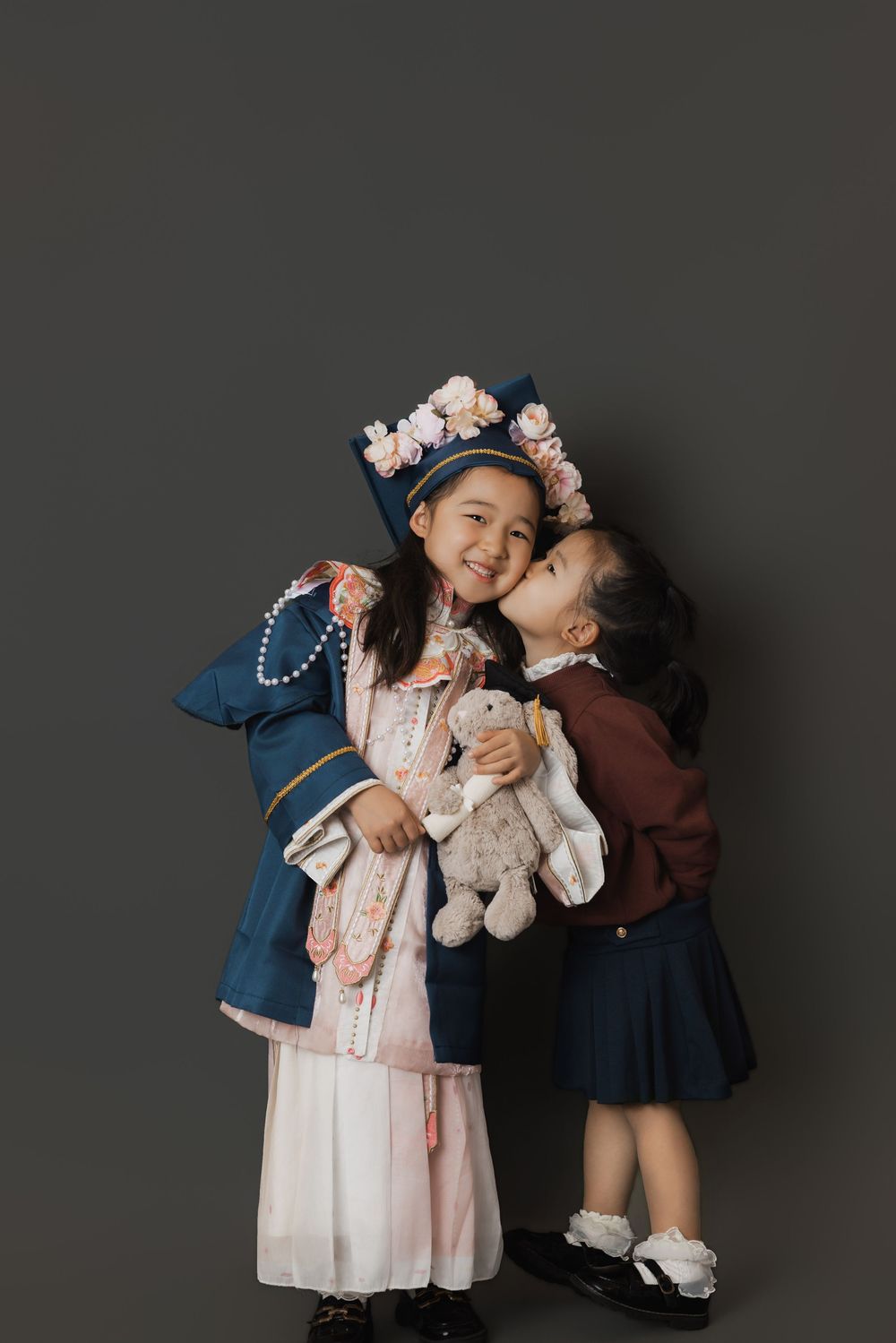 Two young Asian girls in vintage costumes pose together, one kissing the other's cheek while holding a stuffed bunny.