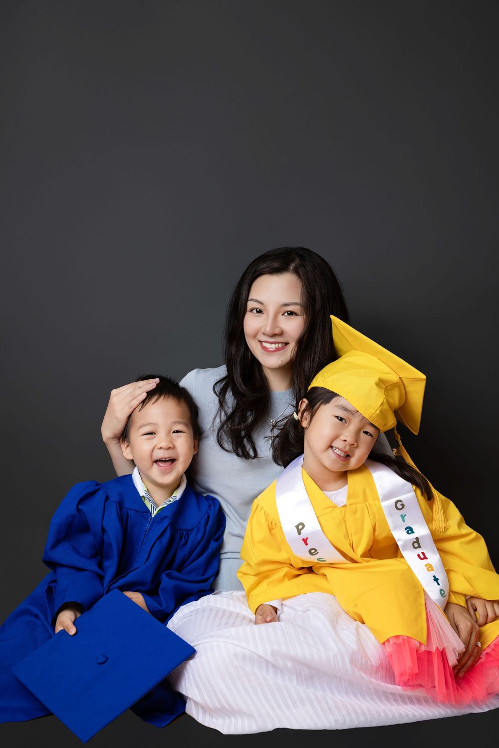 Three people in graduation attire sit together smiling, wearing blue and yellow caps and gowns against a dark background.