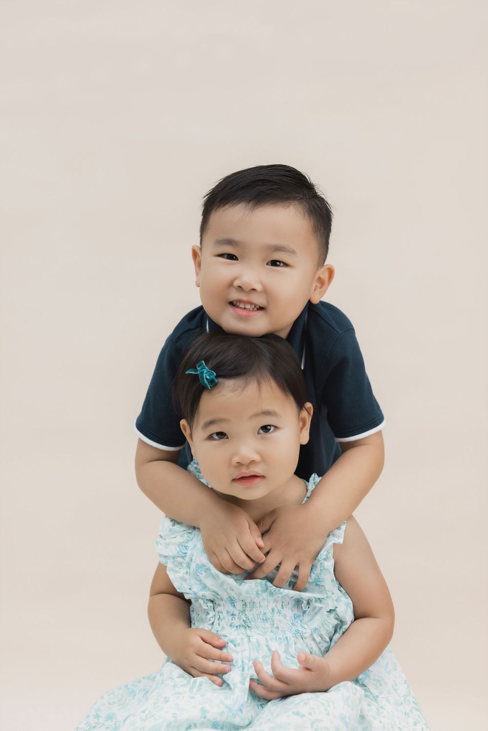 Two young Asian siblings posing together, older boy in navy shirt hugging younger girl in floral dress.