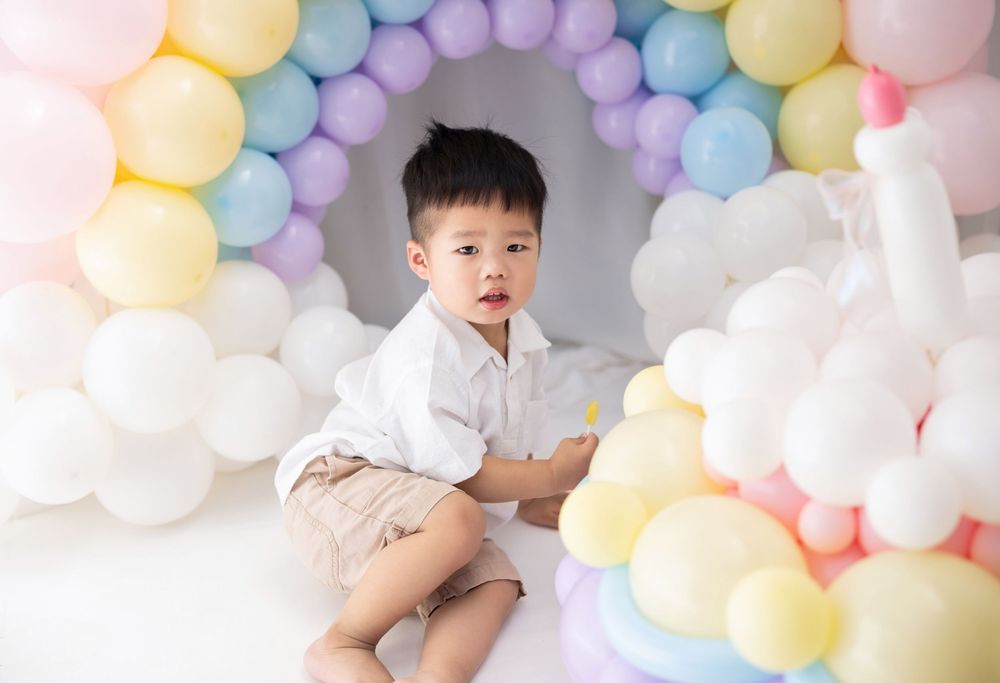Child in white shirt poses against pastel balloon arch backdrop during photo session.